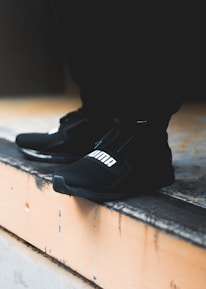 A close-up view of black athletic shoes with a visible brand logo, placed on a concrete step. The shoes are stylishly designed and appear to be suitable for sports or casual wear. The background is blurred, focusing attention on the footwear.