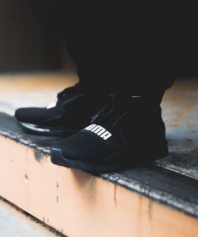 A close-up view of black athletic shoes with a visible brand logo, placed on a concrete step. The shoes are stylishly designed and appear to be suitable for sports or casual wear. The background is blurred, focusing attention on the footwear.
