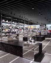 A modern sneaker store with sleek black shelving displaying various athletic shoes and sports apparel. Central seating areas and spotlights provide a contemporary retail ambiance. The signage prominently features a famous sportswear slogan in the background.