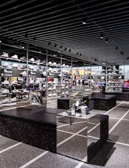 A modern sneaker store with sleek black shelving displaying various athletic shoes and sports apparel. Central seating areas and spotlights provide a contemporary retail ambiance. The signage prominently features a famous sportswear slogan in the background.