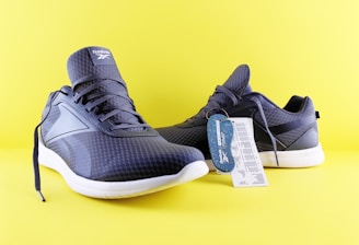 A pair of dark blue athletic shoes with white soles is set against a bright yellow background. The shoes have a textured pattern and display a brand logo on the tongue. There is a tag attached to one of the shoes, providing product information.