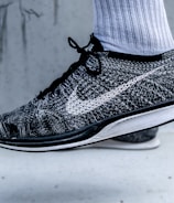 A close-up view of a black and white athletic shoe with a prominent swoosh logo on the side. The shoe is being worn with a white athletic sock, and it appears to be in a mid-air position against a light grey background.