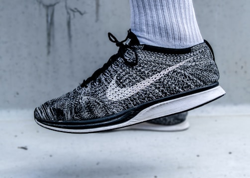 A close-up view of a black and white athletic shoe with a prominent swoosh logo on the side. The shoe is being worn with a white athletic sock, and it appears to be in a mid-air position against a light grey background.