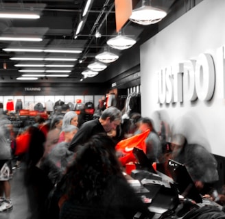 A busy retail store with numerous people examining products and clothing. The store appears to be selling athletic wear, with the phrase 'Just Do It.' prominently displayed on a wall alongside a recognizable orange logo. The interior is well-lit with modern lighting, shelves filled with merchandise, and a counter where people are gathered, possibly checking out or asking questions.