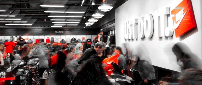 A busy retail store with numerous people examining products and clothing. The store appears to be selling athletic wear, with the phrase 'Just Do It.' prominently displayed on a wall alongside a recognizable orange logo. The interior is well-lit with modern lighting, shelves filled with merchandise, and a counter where people are gathered, possibly checking out or asking questions.