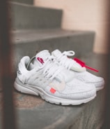 A pair of white athletic shoes with red accents is placed on a set of concrete steps. The shoes have distinct branding and are laced, with one shoe featuring a visible tag.
