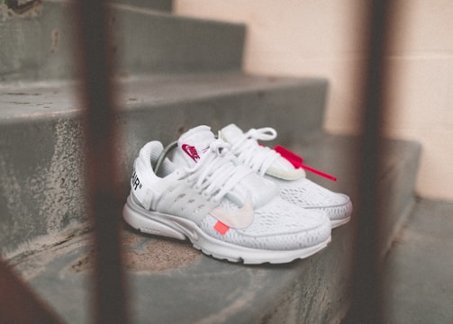 A pair of white athletic shoes with red accents is placed on a set of concrete steps. The shoes have distinct branding and are laced, with one shoe featuring a visible tag.