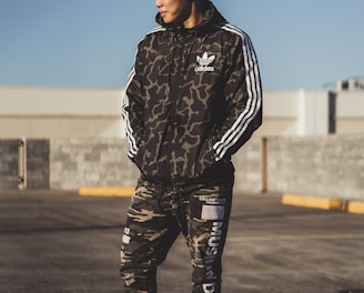 A person stands outdoors wearing a camouflage-patterned athletic outfit with the logo of a well-known sports brand. The individual appears to be in a parking lot or open area under clear daylight. The outfit consists of a jacket with white stripes on the sleeves and matching pants, paired with white sneakers.