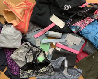 A collection of various athletic apparel and accessories laid out, including sports bras, tops, headbands, and tags. The items are in different colors and patterns, such as pastel pink, blue, and floral designs. A black jacket and several pieces still have brand tags attached.