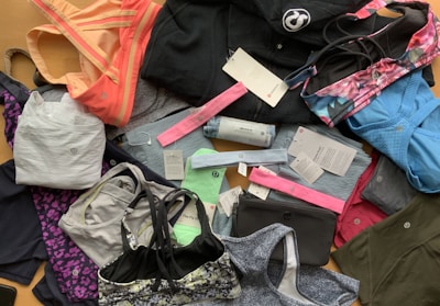 A collection of various athletic apparel and accessories laid out, including sports bras, tops, headbands, and tags. The items are in different colors and patterns, such as pastel pink, blue, and floral designs. A black jacket and several pieces still have brand tags attached.