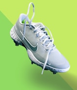 A light blue athletic shoe with a prominent white swoosh symbol and white laces is centered on a vibrant, two-toned green background. The shoe features textured detailing on the sole and upper fabric, indicating it's designed for sports.