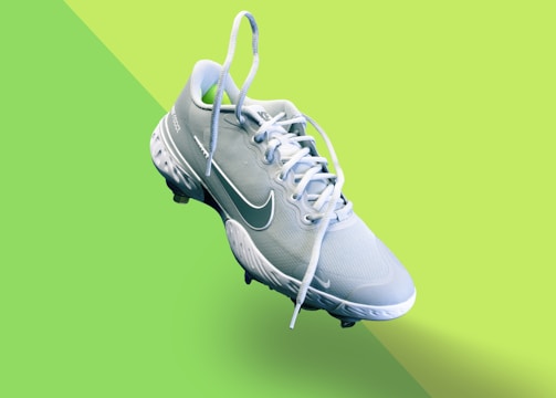 A light blue athletic shoe with a prominent white swoosh symbol and white laces is centered on a vibrant, two-toned green background. The shoe features textured detailing on the sole and upper fabric, indicating it's designed for sports.