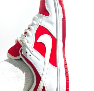 A white and red athletic shoe with a prominent swoosh logo is worn on a foot. The shoe features a lace-up design, primarily white with red accents on the heel, toe, and sole. The fabric appears sleek and clean, with a textured sole for grip.