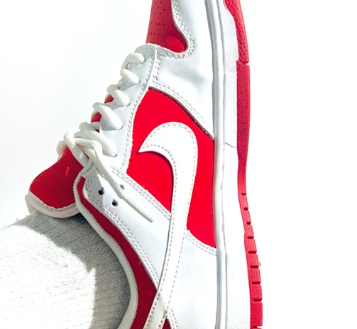 A white and red athletic shoe with a prominent swoosh logo is worn on a foot. The shoe features a lace-up design, primarily white with red accents on the heel, toe, and sole. The fabric appears sleek and clean, with a textured sole for grip.