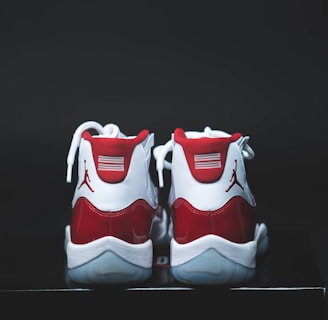 A pair of red and white athletic shoes with a glossy finish, placed on a dark surface. The shoes have white laces and a distinct logo on the heel.