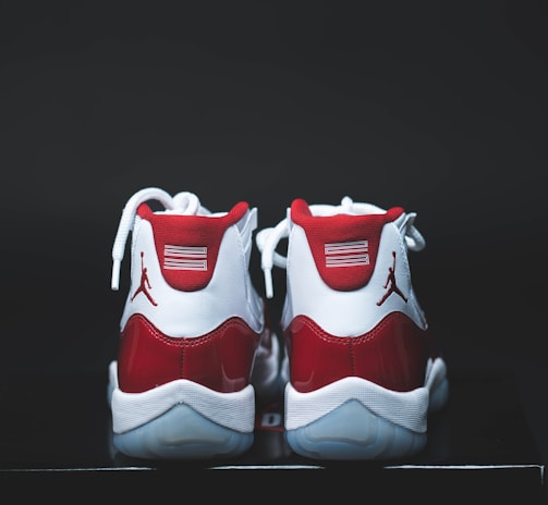 A pair of red and white athletic shoes with a glossy finish, placed on a dark surface. The shoes have white laces and a distinct logo on the heel.