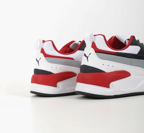 Two athletic shoes with bold red, white, and dark blue color blocking. The shoes feature a sleek, modern design with a flat sole and a recognizable logo in black on the heel. The upper part is made of a mix of textiles with some parts having perforations for breathability.