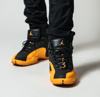 A close-up view of a person wearing black and yellow athletic sneakers. The shoes feature a distinctive brand logo on the tongue and a perforated design. The individual is wearing black pants, and the focus is primarily on the shoes.
