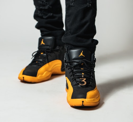 A close-up view of a person wearing black and yellow athletic sneakers. The shoes feature a distinctive brand logo on the tongue and a perforated design. The individual is wearing black pants, and the focus is primarily on the shoes.