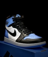 A stylish athletic shoe is placed on a shoebox, featuring a combination of black, white, and light blue. The shoe design includes a prominent logo on the side and a high-top silhouette.