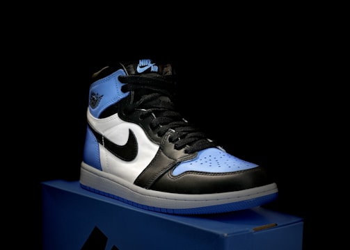 A stylish athletic shoe is placed on a shoebox, featuring a combination of black, white, and light blue. The shoe design includes a prominent logo on the side and a high-top silhouette.