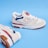 A pair of beige athletic shoes with white laces and blue and red accents. The shoes feature a noticeable logo on the side and are set against a solid blue background.